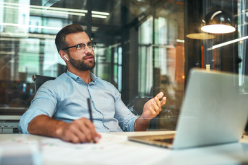 Business talk. Young and successful bearded man in eyeglasses and headphones talking with client by phone while sitting in the modern office