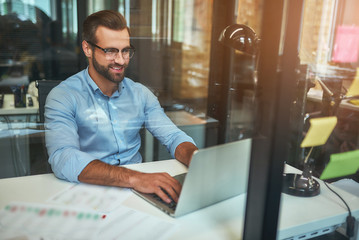 Great results Young bearded businessman in eyeglasses and formal wear looking at laptop and smiling while sitting in the modern office