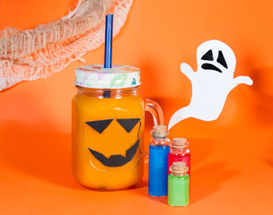  Halloween concept. Pumpkin or carrot drink in a glass jar on an orange background. Nearby are small bottles of red, blue and green drinks. With paper cast in the background.