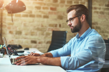 Working day. Side view of young bearded man in eyeglasses and formal wear working on laptop while...