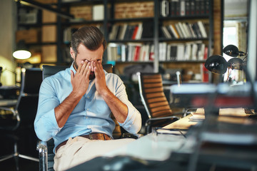 Hard work. Young exhausted bearded businessman covering his eyes and feeling stressed while sitting...