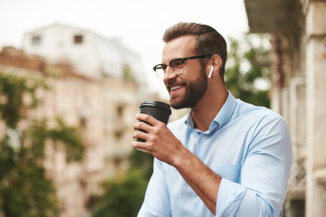 Positive news. Young and handsome bearded man in eyeglasses and headphones holding cup of coffee and talking with friend while standing at the office balcony