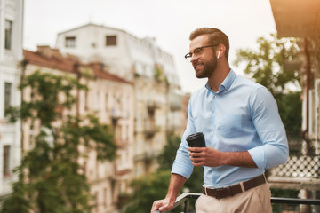 Good news. Young and handsome bearded man in eyeglasses and headphones holding cup of coffee and talking with friend while standing at the office balcony - obrazy, fototapety, plakaty