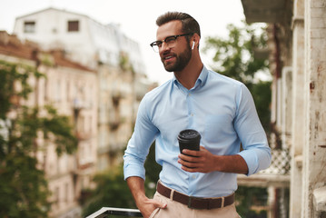 Nice talk. Young and handsome bearded man in eyeglasses and headphones holding cup of coffee and talking with friend while standing at the office balcony