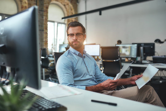 Checking The Reports. Busy Bearded Man In Eyeglasses And Formal Wear Holding Documents And Looking At Computer Monitor While Sitting In The Modern Office