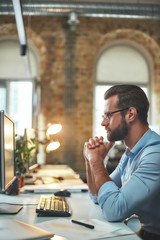 The best workplace. Side view of young and successful bearded man in eyeglasses and formal wear looking at computer monitor during his working day in modern office