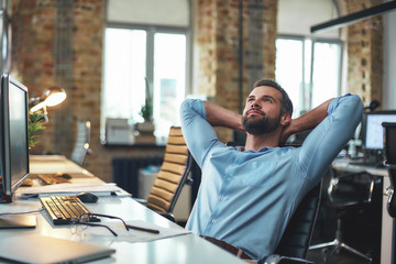 Enjoying break. Satisfied young bearded businessman leaning back with hands behind head and relaxing while sitting in the modern office - obrazy, fototapety, plakaty