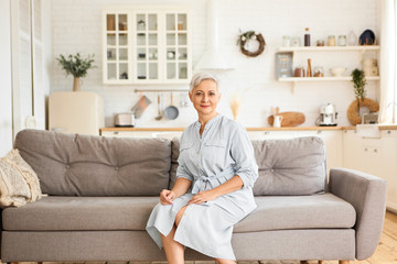 Indoor image of attractive elegant Caucasian retired woman with short gray hairdo wearing stylish blue dress sitting on sofa in relaxed pose, looking at camera with calm joyful smile. People and age
