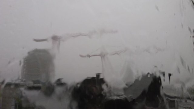 Starker Regen rinnt w&auml;hrend eines Gewitters an einer Fensterscheibe herunter wie ein kleiner Bach mit sch&ouml;nen Tropfen und Prasseln und im Hintergrund zwei Baukr&auml;nen einer Baustelle