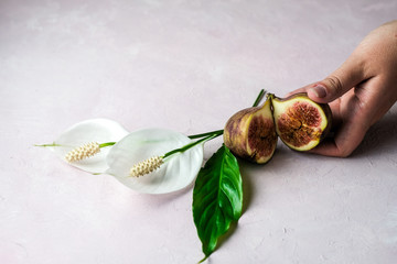 Hand holding a fresh whole and cut figs with some flowers on a light pink marbled background, copy space. Selected focus