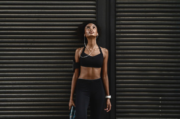 Front view of sporty woman listening music on earphones and leaning to a wall outdoors taking break after workout