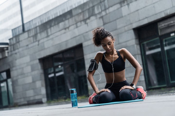 Young fitness woman in earphones sitting relaxed on yoga mat after workout with a water bottle...