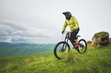 Front view of a man on a mountain bike standing on a rocky terrain and looking down against a gray...