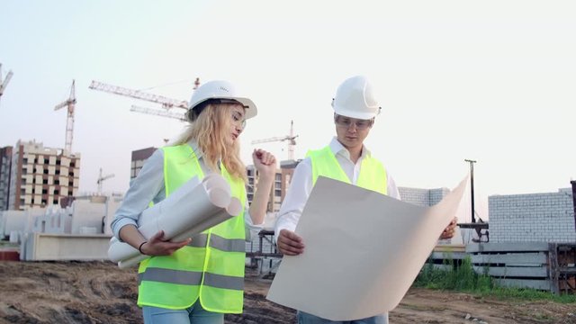 Workers with drawings at the construction site. Two workers man and woman in protective harhats working with drawings at the construction site outdoors