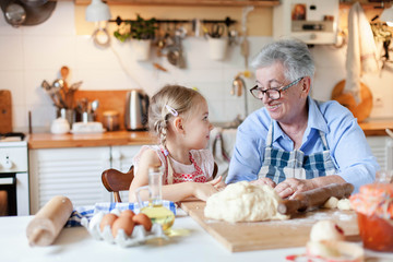Senior woman and child make dough for bread together. Family is cooking in cozy kitchen at home....