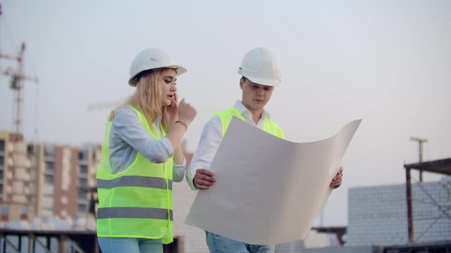 Workers with drawings at the construction site. Two workers man and woman in protective harhats working with drawings at the construction site outdoors