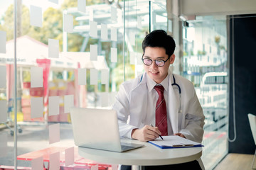 Male doctor writing on clipboard and looking laptop in the office.