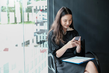 Asian woman who is sitting smiling and using a mobile phone while waiting for a job interview. Job application concept