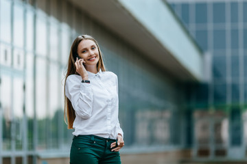 Fototapeta premium Young famale calling on a smartphone. Attractive businesswoman talking by phone. Office building Background. Technology internet and happy people concept.