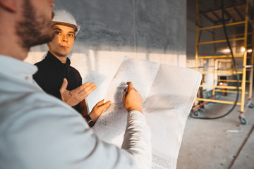 Fototapeta premium Building engineer and worker hold blueprint in hand while discussong construction site. Industrial engineers have meeting on build
