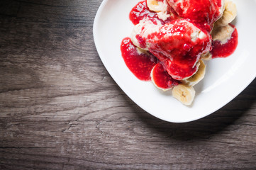 Closeup Vanilla ice cream poured with strawberry or raspberry jam with sliced banana slices on a white plate on a dark wooden table. You can do this at home on vacation