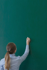 Rear view of a student or teacher with long brunette hair writing on a blank green blackboard or...
