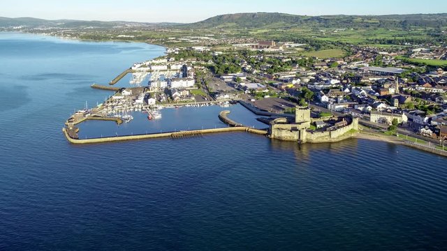 Belfast Lough with Medieval Norman Castle in Carrickfergus near Belfast in sunrise light. Aerial 4K ideo with marina, breakwater, yachts, parking, town and far view of Belfast in the background