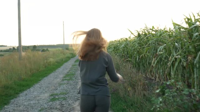 Flirty red-haired girl smiles and runs away along country road near a corn field. Beautiful young woman in khaki sport suit running outdoors at sunset. Agricultural Footage. 