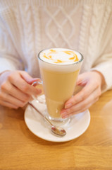 A glass of coffee with cream and foam in the hands of a girl in a white knitted sweater. Cozy lovely winter warm picture.