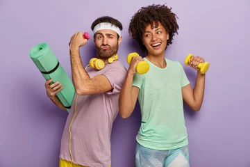 Tafelkleed Glad multiethnic husband and wife attend sport center, exercise with dumbbells, hold fitness mat, stand back to each other, have funny happy looks, wear t shirts, isolated on purple background © Wayhome Studio