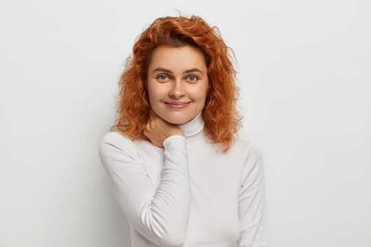 Headshot of pleasant looking ginger woman with satisfied expression, touches neck, has natural beauty, smooth healthy skin, wears white turtleneck sweater, models in studio, feels delighted.