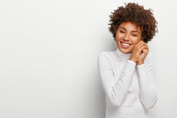 Fototapeta na wymiar Joyous black woman with pleasant smile, keeps hands together near face, looks happily, dressed in casual clothing, expresses good emotions, poses against white wall, blank space for advertisement