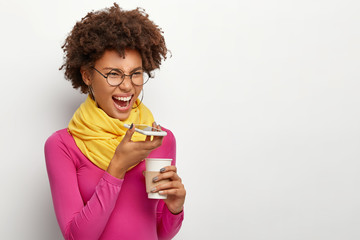 Annoyed woman shouts at speaker of cell phone, makes voice call, angry with friend for not coming on meetig, drinks hot aromatic coffee, wears spectacles for vison correction, colored outfit