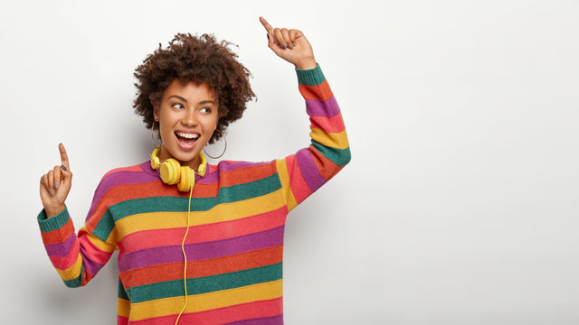 Energetic carefree African American female dances with raised arms, enjoys nice track, listens mucis via headset, wears casual loose striped colorful jumper, feels relaxed and glad, isolated on white