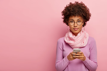 Beautiful young woman with curly dark hair, stays in touch, uses modern gadget, makes her own blog, connected to wireless internet, wears glasses, big hoop earrings, scarf and turtleneck poses on pink - Powered by Adobe
