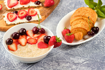 white bowl of porridge with strawberries and cherries, croissants and crispbread with cheese and strawberries on a gray background