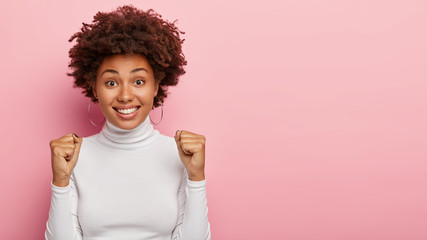 Studio shot of attractive positive woman raises clenched fists, anticipates for some results, has pleasant smile, rejoices something, dressed in white casual jumper, isolated on pink background