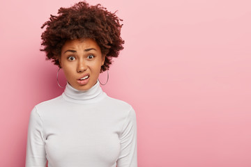 Discontent woman with Afro haircut, smirks face, sees something unpleasant, wears hoop earrings, casual white jumper, isolated on pink wall with blank space. Negative emotions and facial expressions