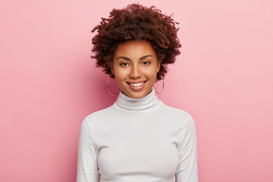 Gorgeous lovely woman has pleasant smile on face, healthy skin, Afro haircut, wears casual white turtleneck jumper, round earrings, looks happily directly at camera, has no make up, feels joyful