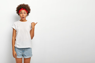 Impressed astonished young dark skinned woman points tumb right, shows blank white copy space,...