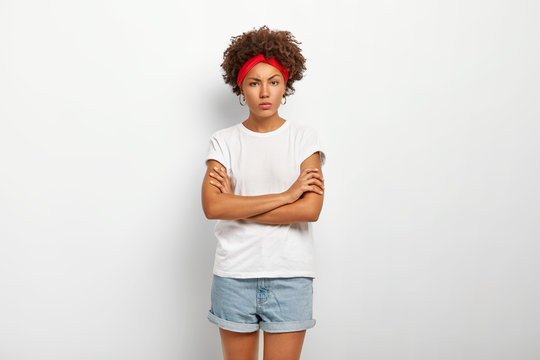 Insulted angry curly woman frowns face, stands with hands crossed over chest, makes defensive pose, wears white t shirt, jean shorts, expresses hate and distrust, models against white background