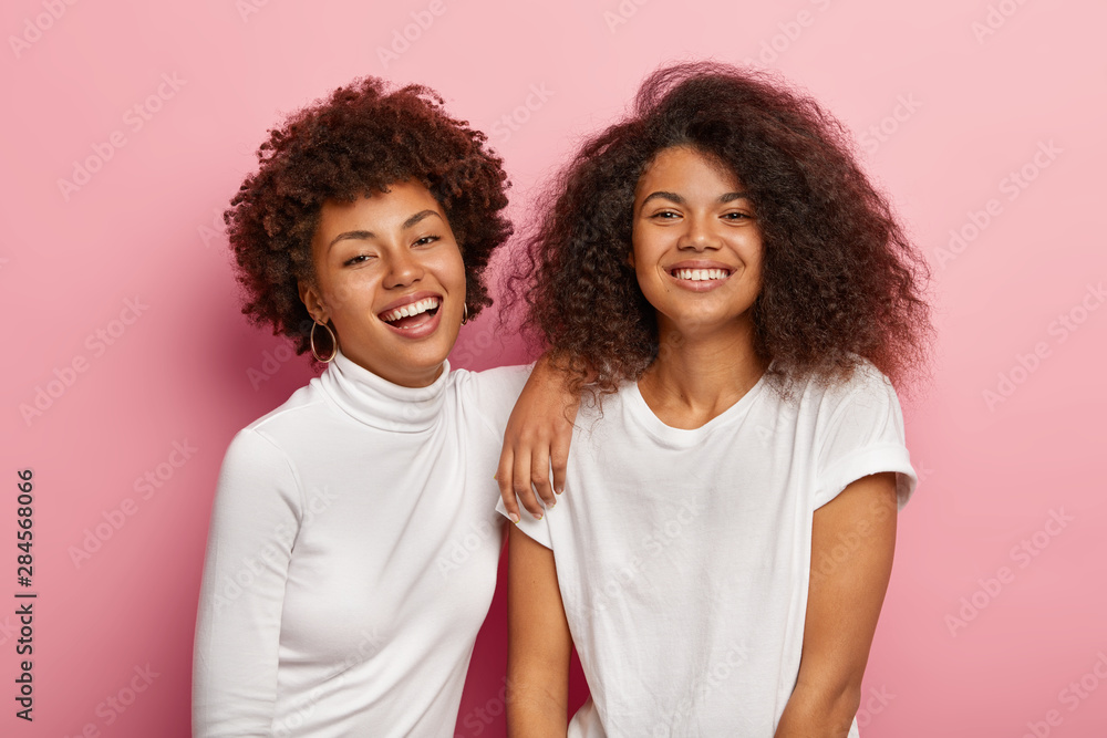 Wall mural Lovely happy dark skinned women have fun together, enjoy spare time, smile happily, have natural curly hair, dressed casually, isolated on pink background. People, youth and emotions concept