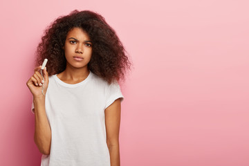Unhappy curly haired woman holds clean cotton tampon, sad to have menstruation, cares about hygiene protection during periods, wears white t shirt, stands over pink background, copy space area