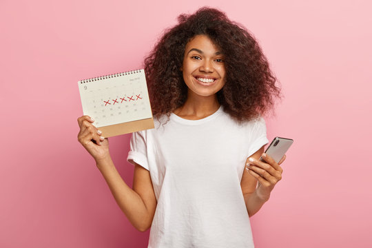 Happy curly woman holds periods calendar, checks menstruation days on mobile phone application, cares about women health, dressed casually, isolated on pink studio wall. Pms and wellness concept