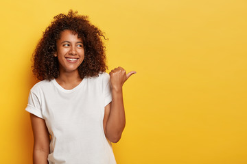 Adorable female student with bushy curly hair points thumb right, feels happy and relaxed, wears...