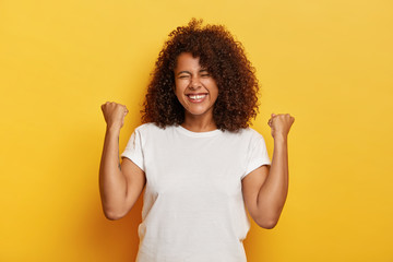 Isolated shot of beautiful successful woman with curly hair, raises clenched fists, celebrates...
