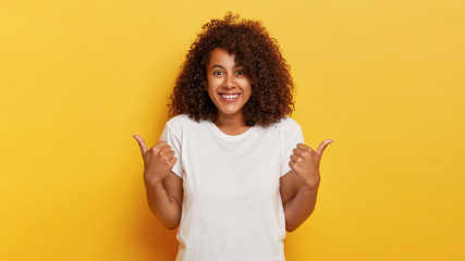Happy curly haired girl makes thumbs up sign, demonstrates support and respect to someone, smiles pleasantly at camera, achieves desirable goal, wears white t shirt, isolated on yellow background