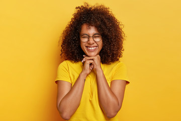 Pretty charming Afro girl with curly hair, rejoices life, keeps hands under chin, feels overjoyed and satisfied, has natural appearance, wears bright yellow t shirt, poses indoor. Happiness concept