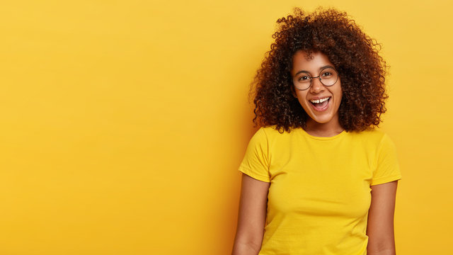 Upbeat Glad Curly Haired Adorable Girl Laughs Out From Happiness, Rejoices Pleasant Moments In Life, Has Appealing Look, Wears Big Transparent Glasses And Casual Yellow T Shirt, Feels Joyous