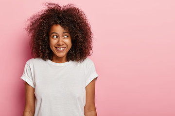 Attractive dark haired curly female looks aside, has charming smile, being in good mood after date or party, wears casual mockup t shirt, stands over rosy wall, free space aside. People and emotions